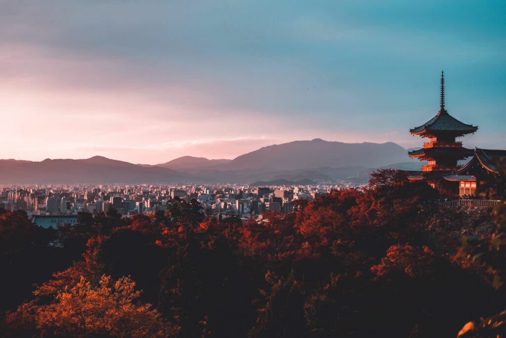 Pagoda in Japan overlooking orange and red trees in the foreground with mountains overlooking city buildings in the background. Photo by Su San Lee on Unsplash.